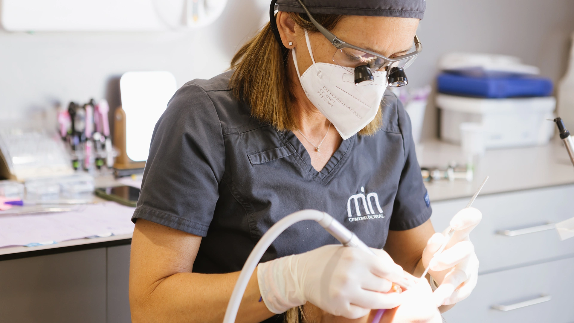 Professional de MiN Centre Dental atenent una pacient amb instrumental dental i ulleres d’augment.